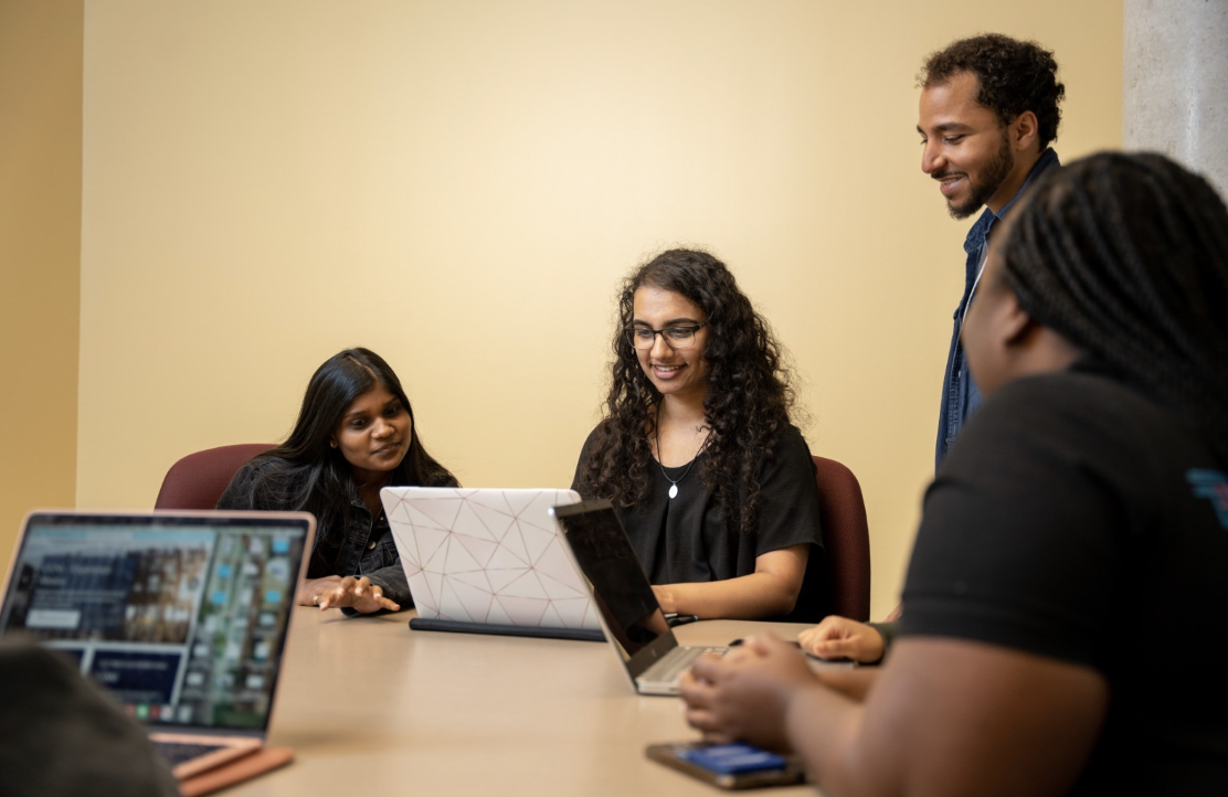 Photo of three people sitting around a table, peering over a laptop smiling. A third and fourth student seated in the lower left and right corner, and one has their own laptop.