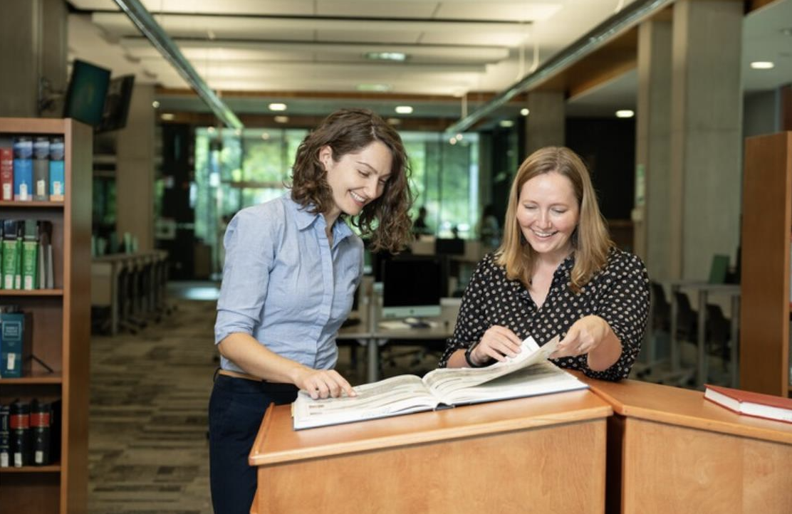 Photo of two caucasian women standing at a lecturn in a library looking at an open book and smiling. 