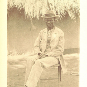 An archival image of a Black man sitting in a tan suit and fedora from the 1930s.