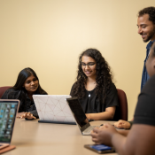 Photo of three people sitting around a table, peering over a laptop smiling. A third and fourth student seated in the lower left and right corner, and one has their own laptop.