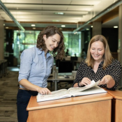 Photo of two caucasian women standing at a lecturn in a library looking at an open book and smiling. 