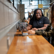 Two students sit at a counter height wooden table, each with a laptop. They are smiling at each other int he UTSC Library.
