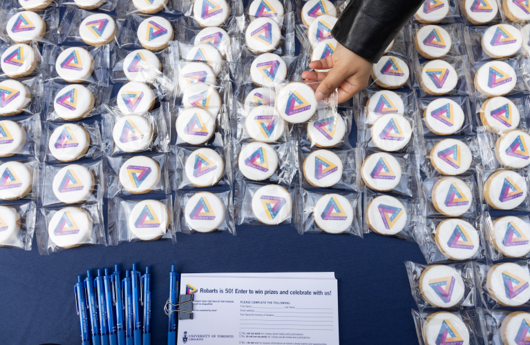 white cookies on a table with a blue, yellow and fuchsia gradient triangle with the words "ROBARTS FIFTY" cover a blue table cloth. A hand grabs one cookie.